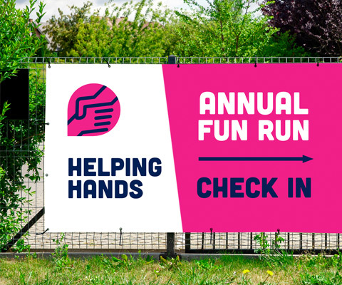 A large outdoor helping hands banner with a pink background that reads Annual fun run check in sits on a fence in front of large trees.