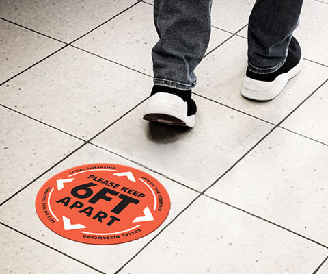 A circular red floor decal that reads "Please keep 6ft apart" with arrows with the feet of a person walking by.